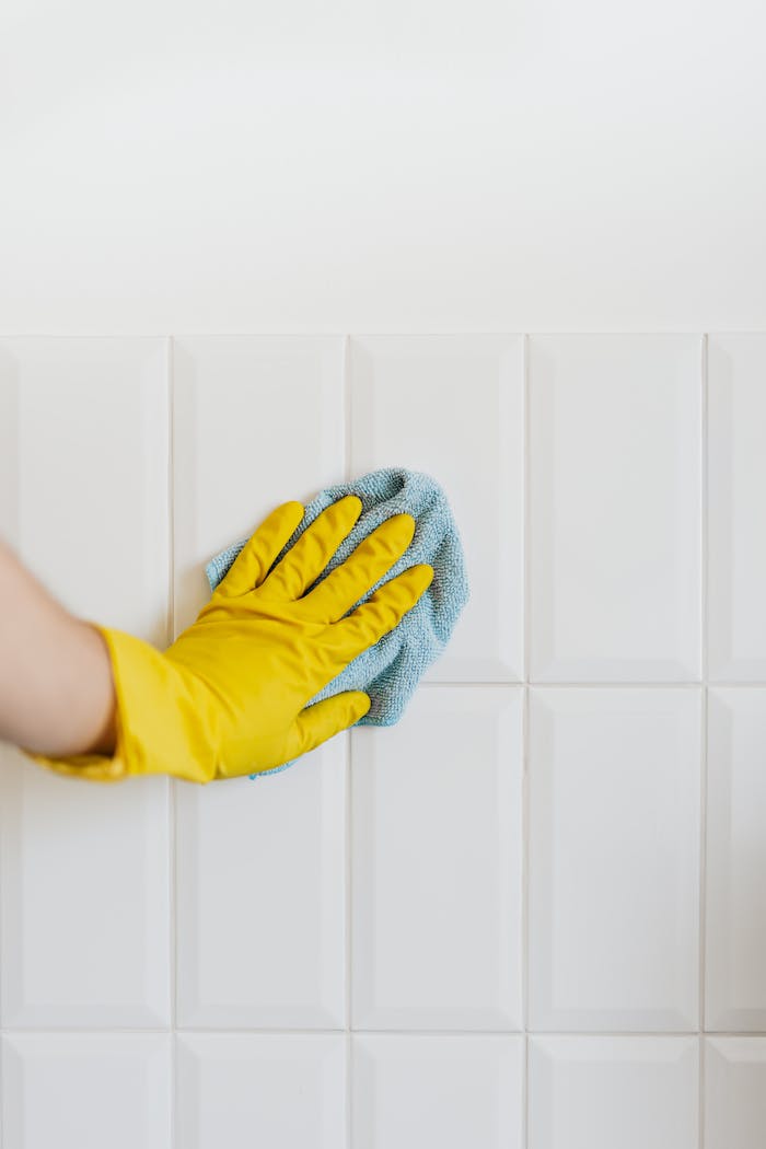 Close-up of a gloved hand wiping a white tiled wall with a blue cloth, symbolizing cleanliness and hygiene.